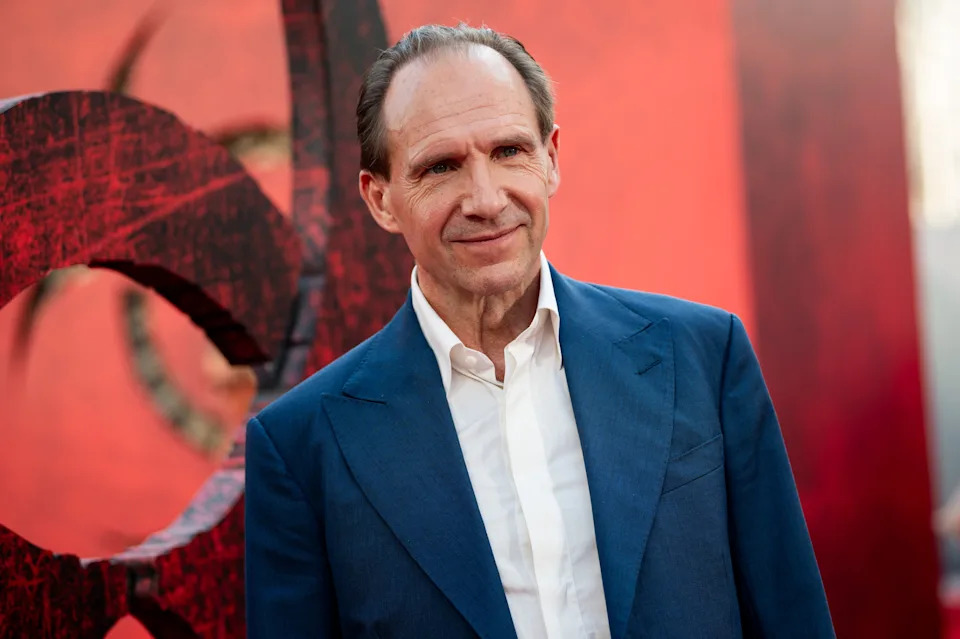 Person in a blue suit and white shirt at an event, standing against a dramatic red backdrop, smiling slightly