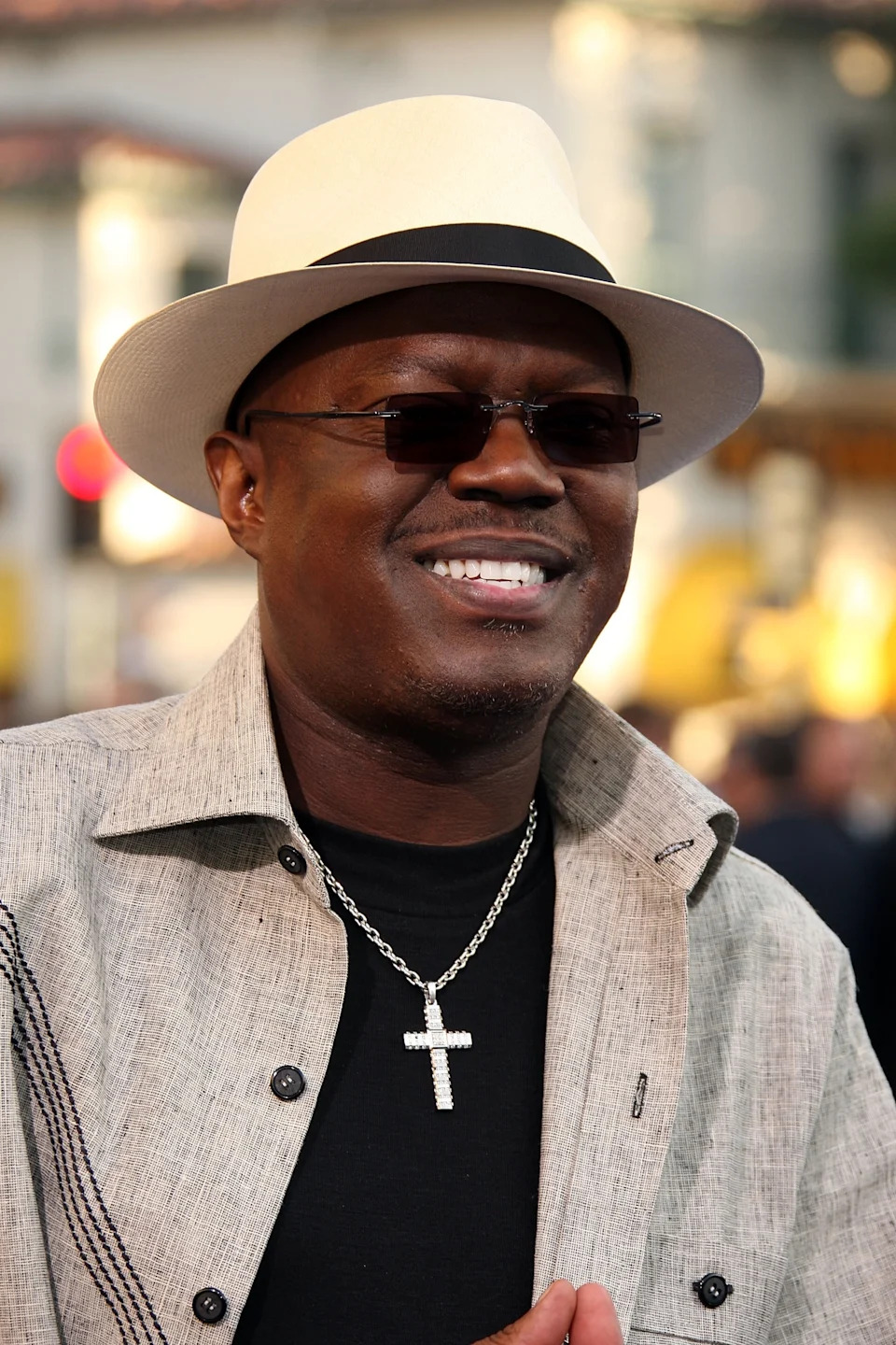 Bernie Mac in a stylish hat and sunglasses, wearing a cross necklace and a light-colored textured jacket, smiling outdoors