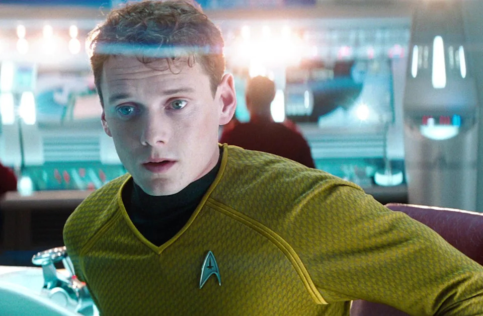 Anton Yelchin wearing a Starfleet uniform is on a futuristic spaceship bridge, appearing surprised or concerned