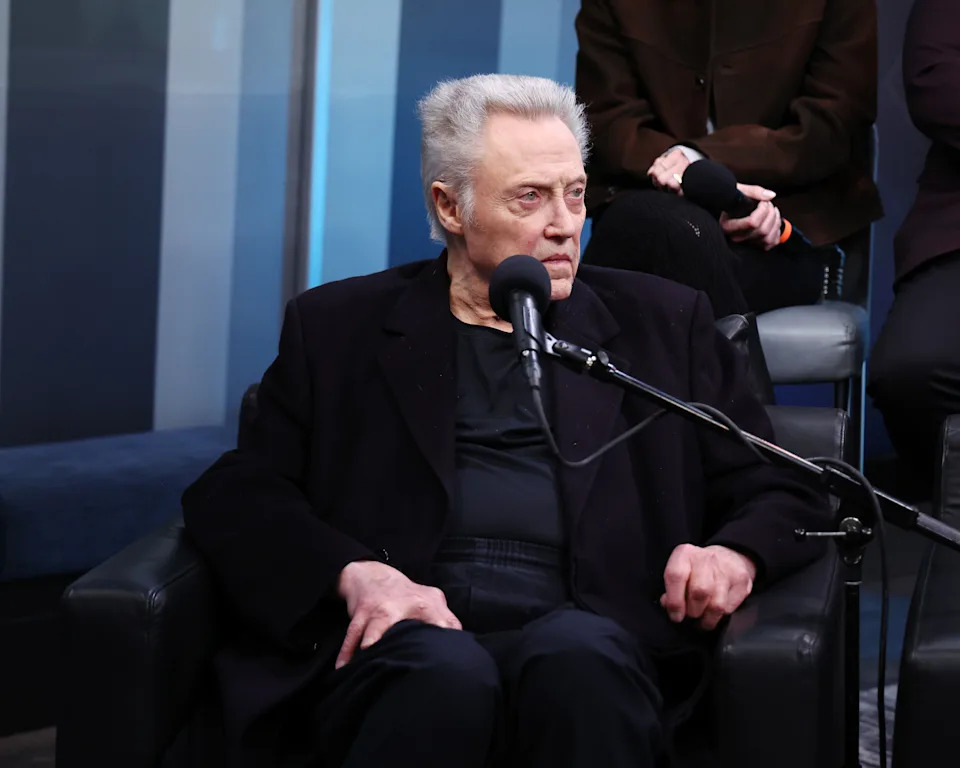 A man sits on a chair speaking into a microphone, wearing a dark jacket and shirt. Another person is seated in the background