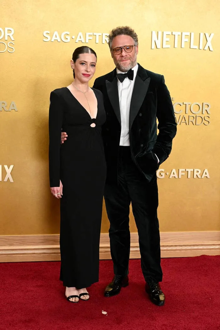 Two people on a red carpet; one in a black gown with a long train, the other in a velvet suit with a bow tie