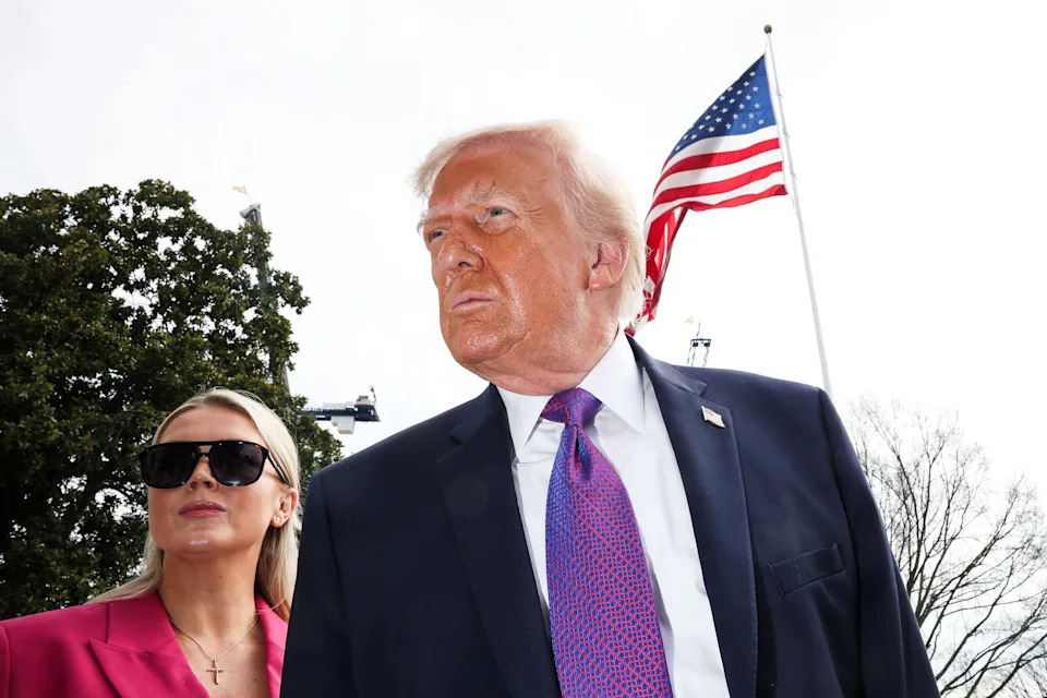 Two individuals outdoors, one in a suit and tie, with an American flag in the background