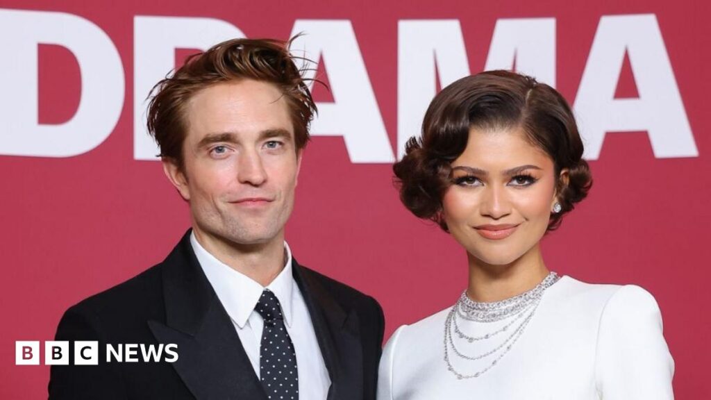 Robert Pattinson and Zendaya smiling at the camera. He has brown hair and is wearing a black suit jacket, black tie with white polka dots and a white shirt. Zendaya is wearing a white dress and diamond earrings and necklaces. She has short brown hair.