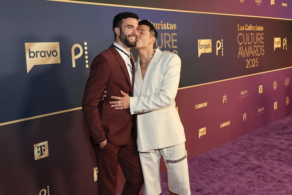 Two individuals on a red carpet; one in a burgundy suit and the other in a white suit, smiling and embracing at a culture awards event