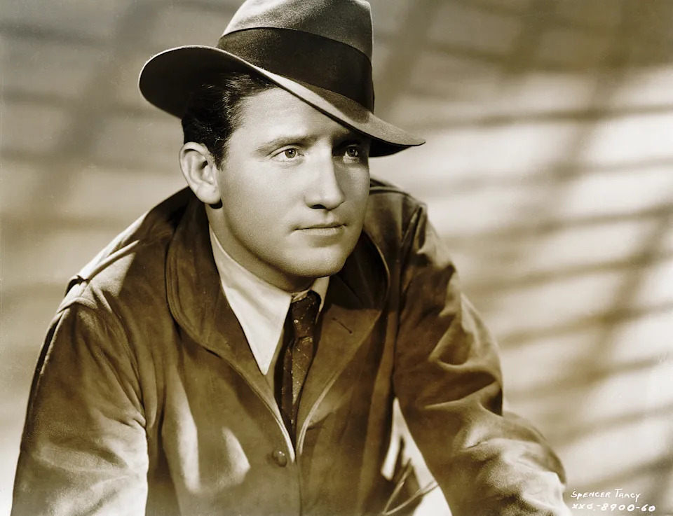 Spencer Tracey in a fedora and jacket, sitting pensively