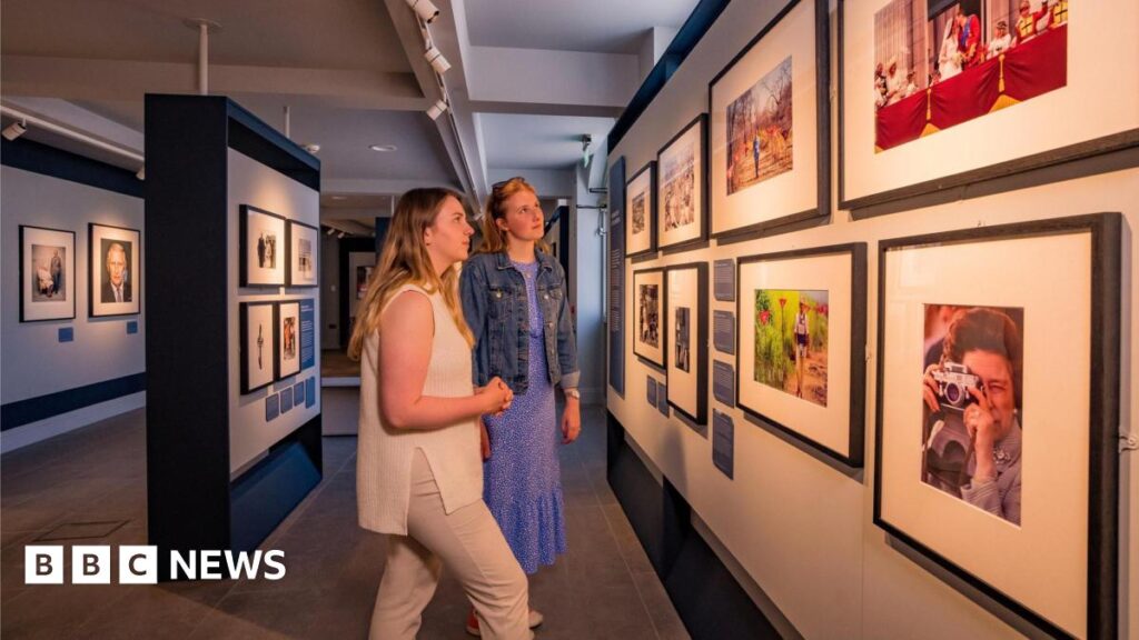 Two women with blonde hair in an art gallery looking at photographs of the British royal family on a wall.