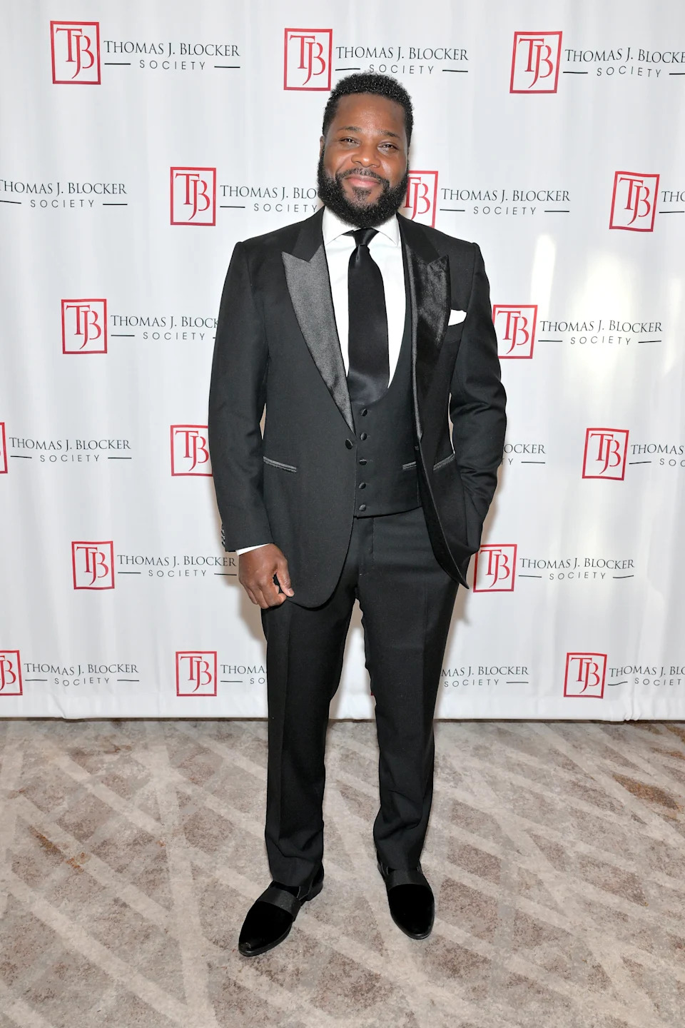 Malcolm Jamal Warner in a tuxedo at an event, standing in front of a backdrop with logos
