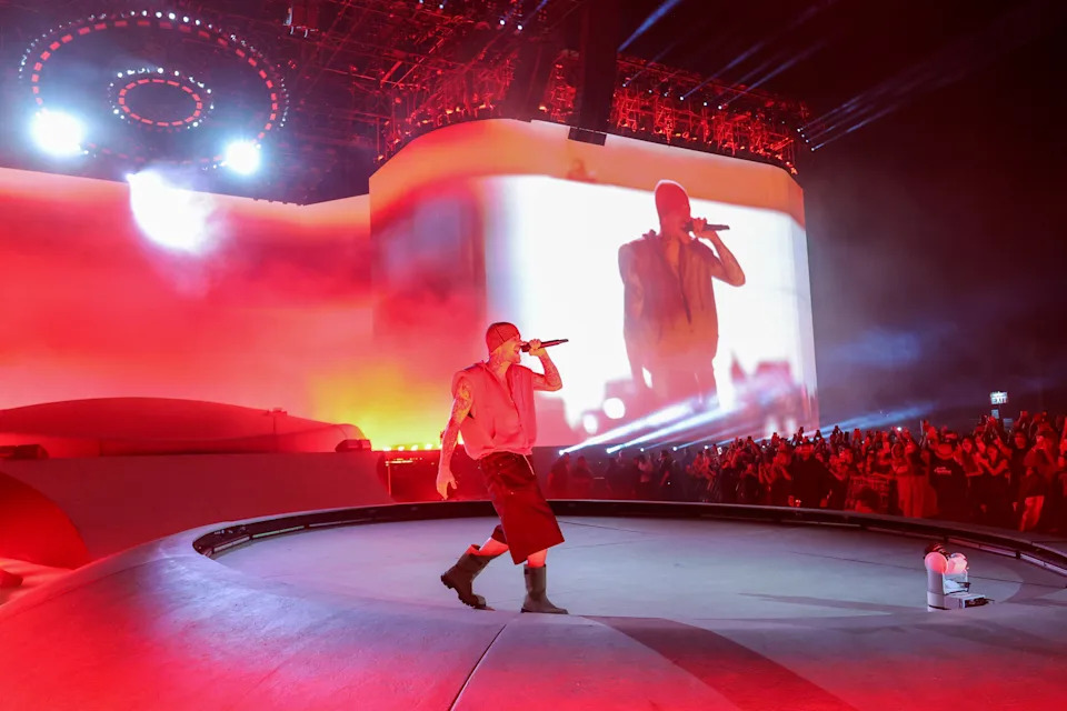Justin Bieber on stage holding a microphone, with a large screen showing their image in the background, surrounded by a vibrant concert atmosphere