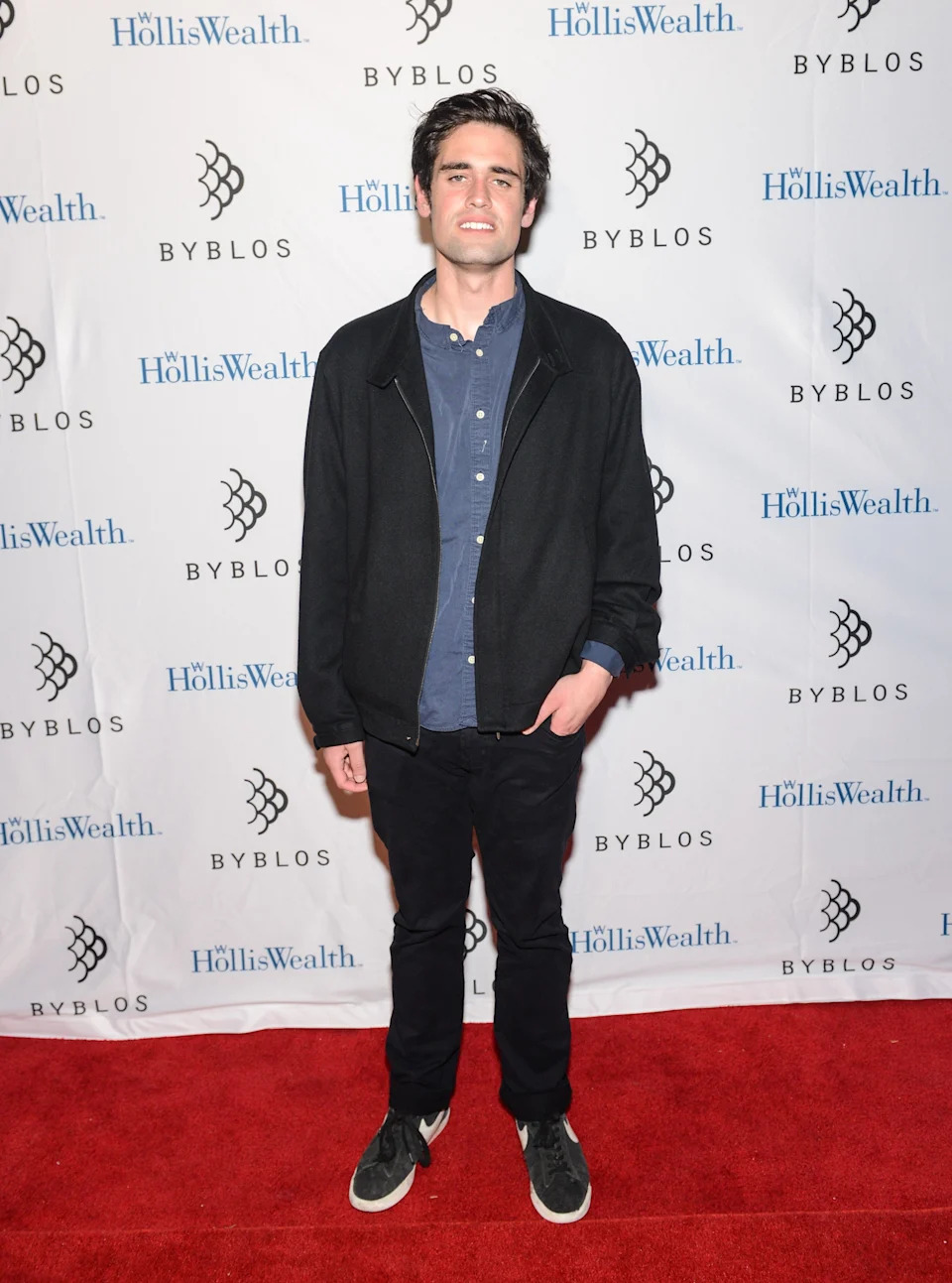 Nick Reiner on red carpet wearing a casual outfit with a jacket, shirt, and pants, standing in front of a backdrop with logos