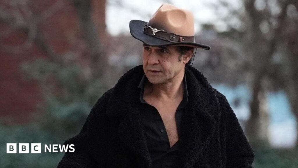Chico arriving at court, wearing a brown cowboy hat with a silver cattle head adornment. He wears a black furry coat with a black open shirt underneath