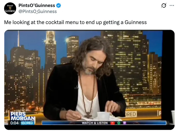 A person with long hair and a beard, wearing a deep V-neck shirt and blazer, looks at papers on a talk show set with a cityscape backdrop