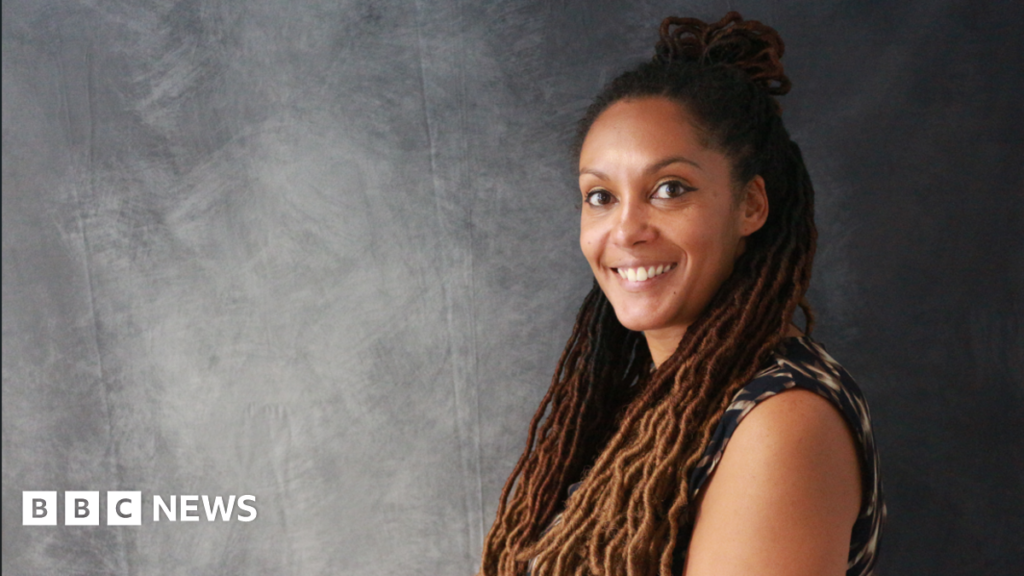 A woman with long braided afro hair is smiling as she is standing next to a grey backdrop.