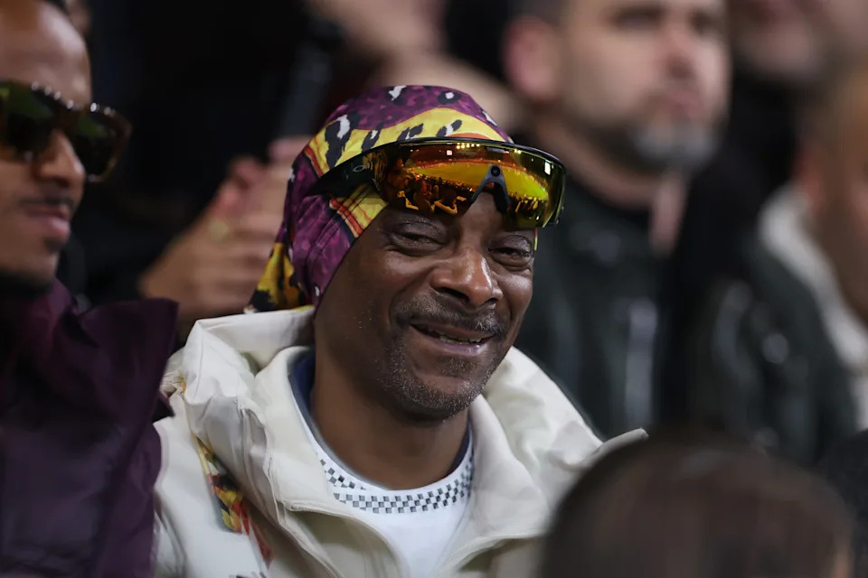 Snoop Dogg wearing a headscarf and sunglasses, smiling while seated among a crowd at an event