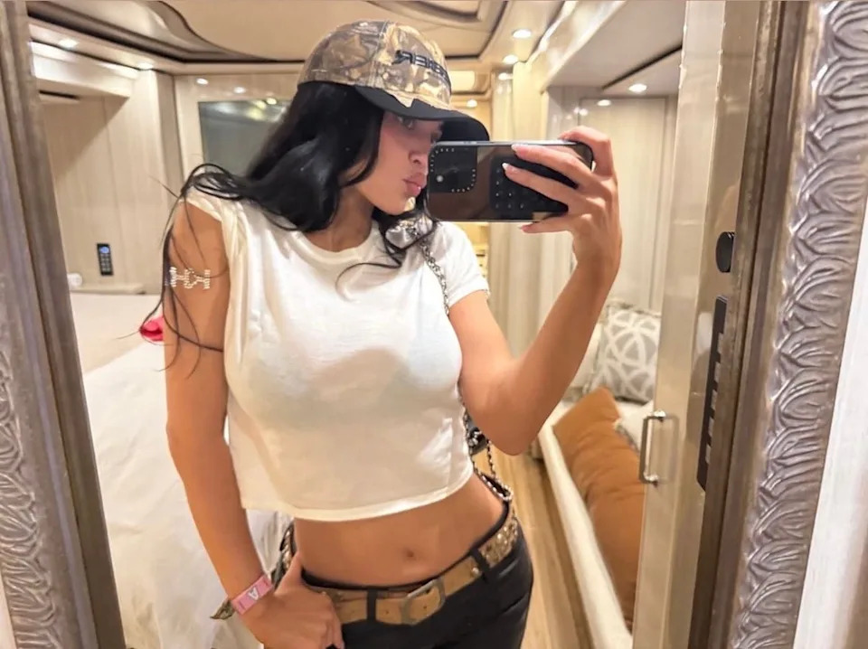 Kylie Jenner taking a mirror selfie wearing a white crop top, camo hat, and belt with decorative skulls, posing in a well-lit room