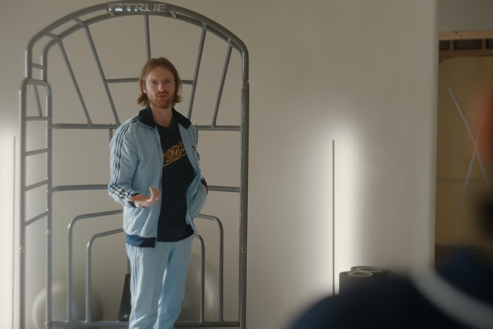 Finneas stands next to a workout frame wearing a tracksuit in a scene from Beef season 2