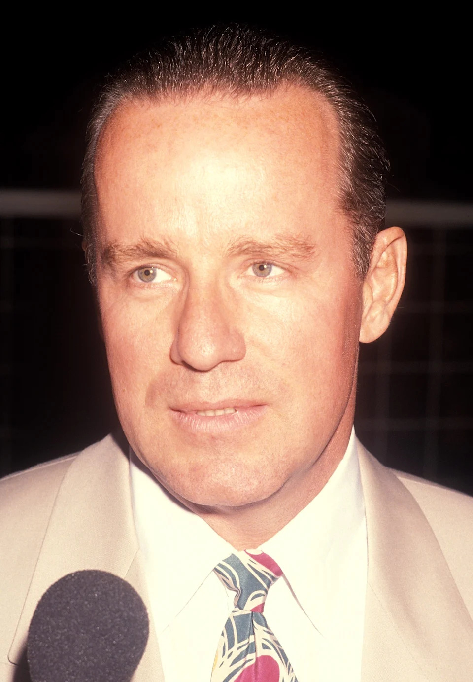 Phil Hartman in a suit and abstract-patterned tie speaks into a microphone, with a serious expression