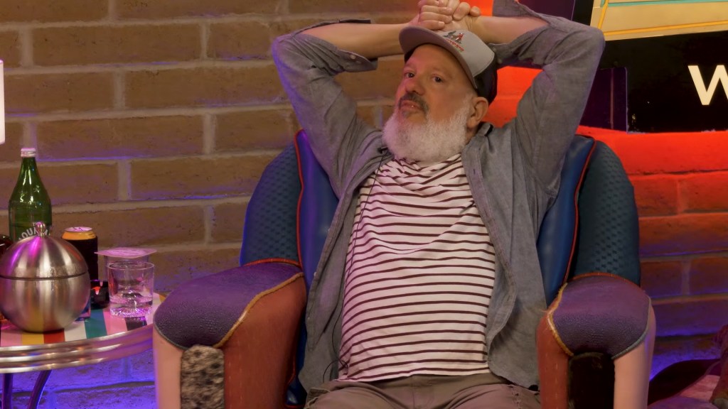 David Cross sitting in a chair, with his hands behind his head.