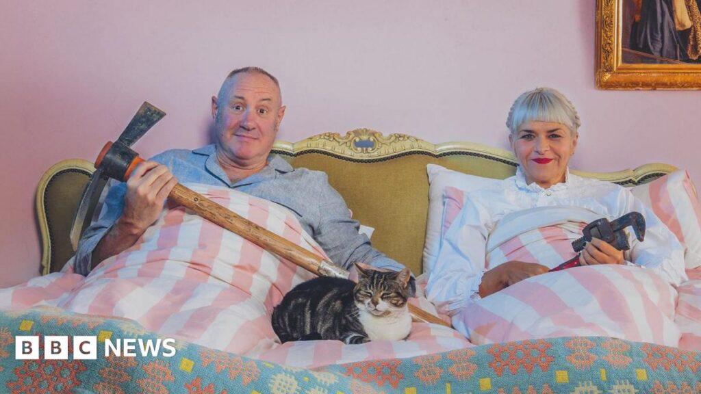 Keith and Marj are sat up in a bed with pink and white striped bedding. There is a cat sat between them. Both of them are holding building tools. The wall behind them is pink and has a hole in it exposing wooden panels. On the same wall is a gold mirror.
