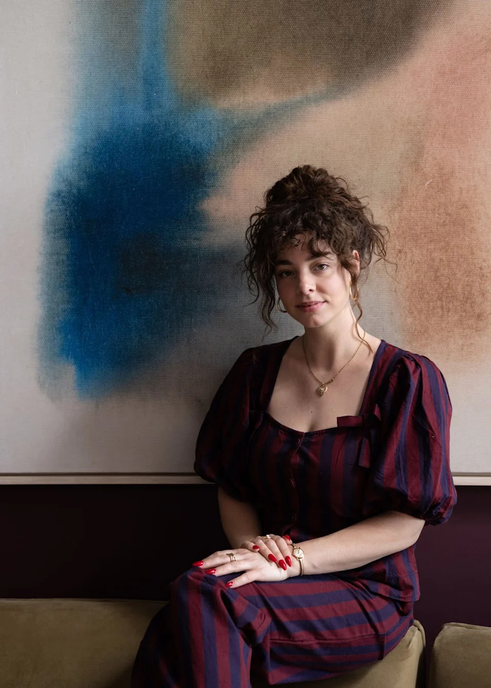 A seated individual in stylish attire next to an abstract painting.