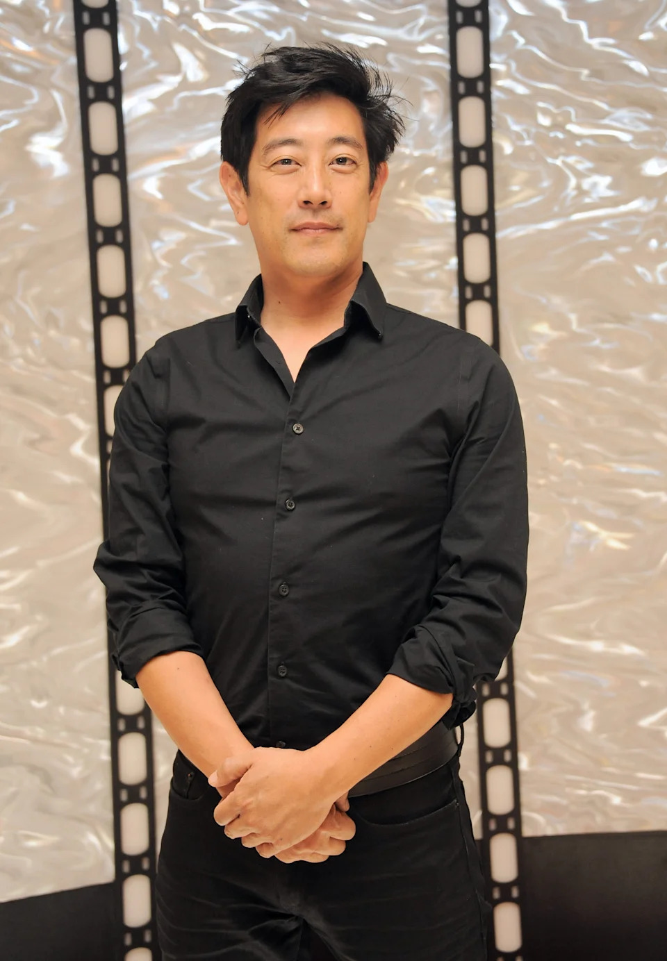 Grant Imahara standing against a patterned wall, wearing a button-up shirt, looking confidently at the camera