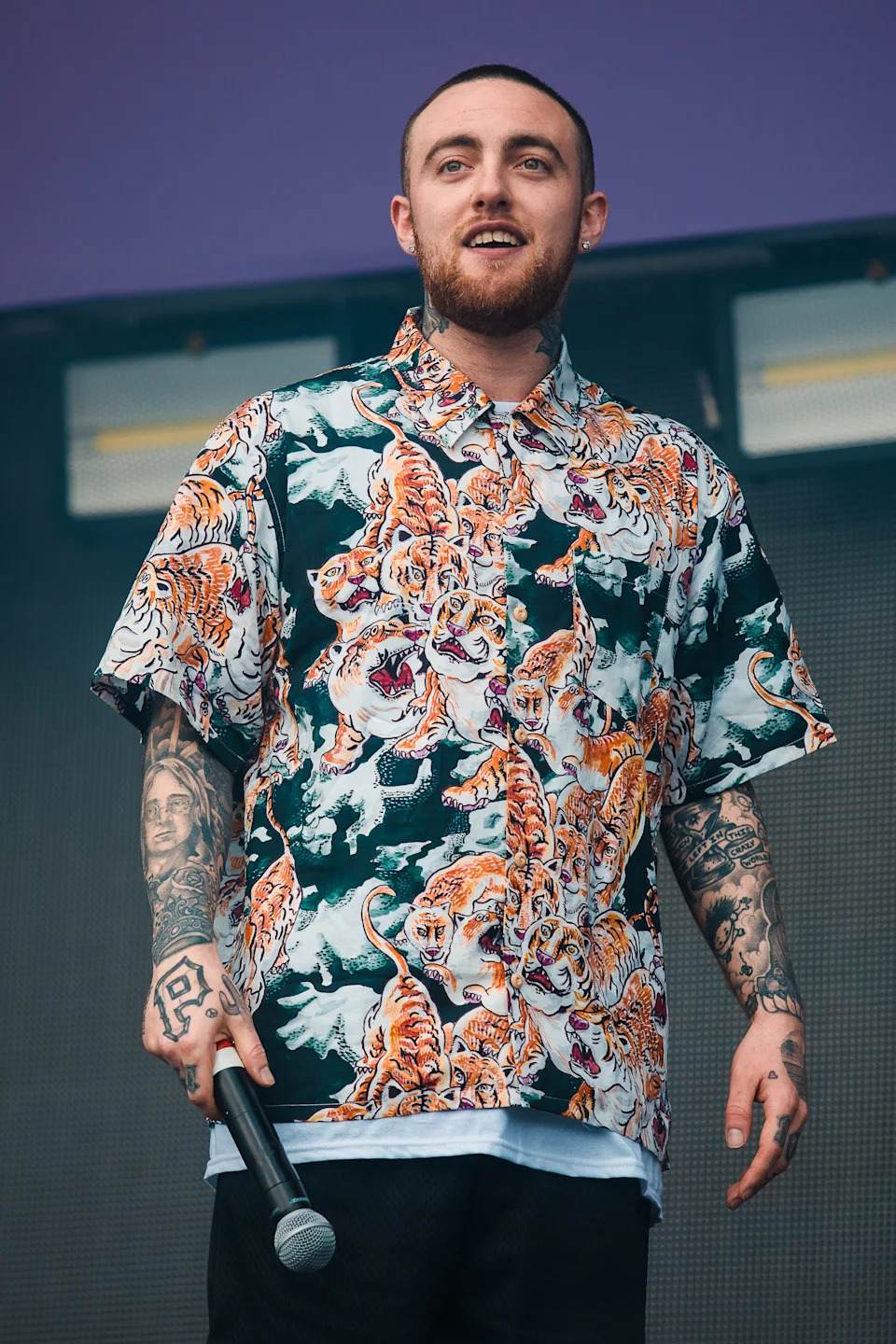 Person in a tiger-print shirt holding a microphone, standing confidently on stage with tattoos visible on arms