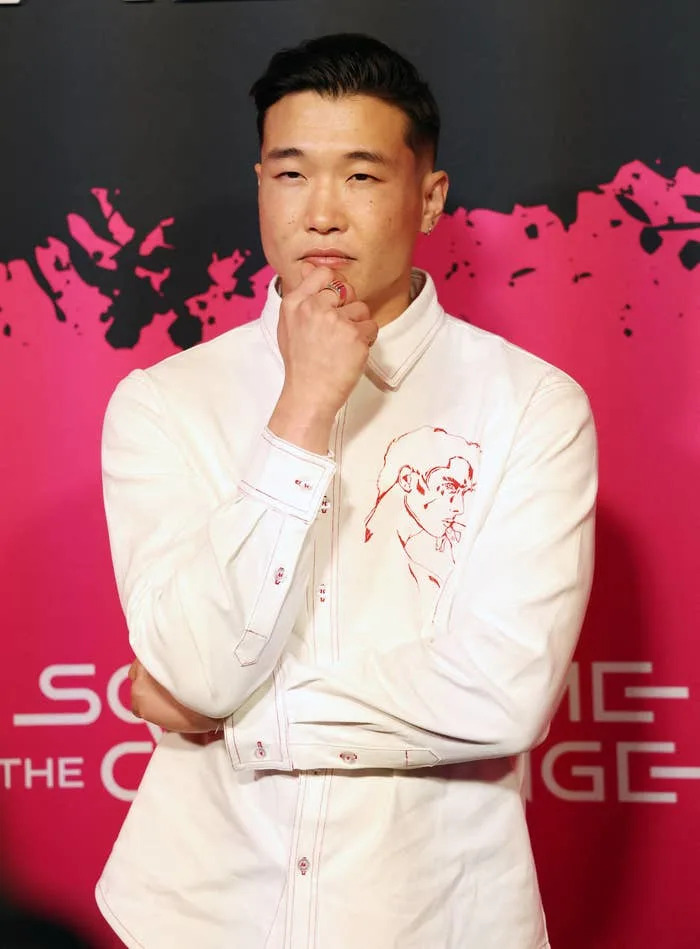 Person posing on a red carpet in a white shirt with an artistic design. They have a thoughtful expression
