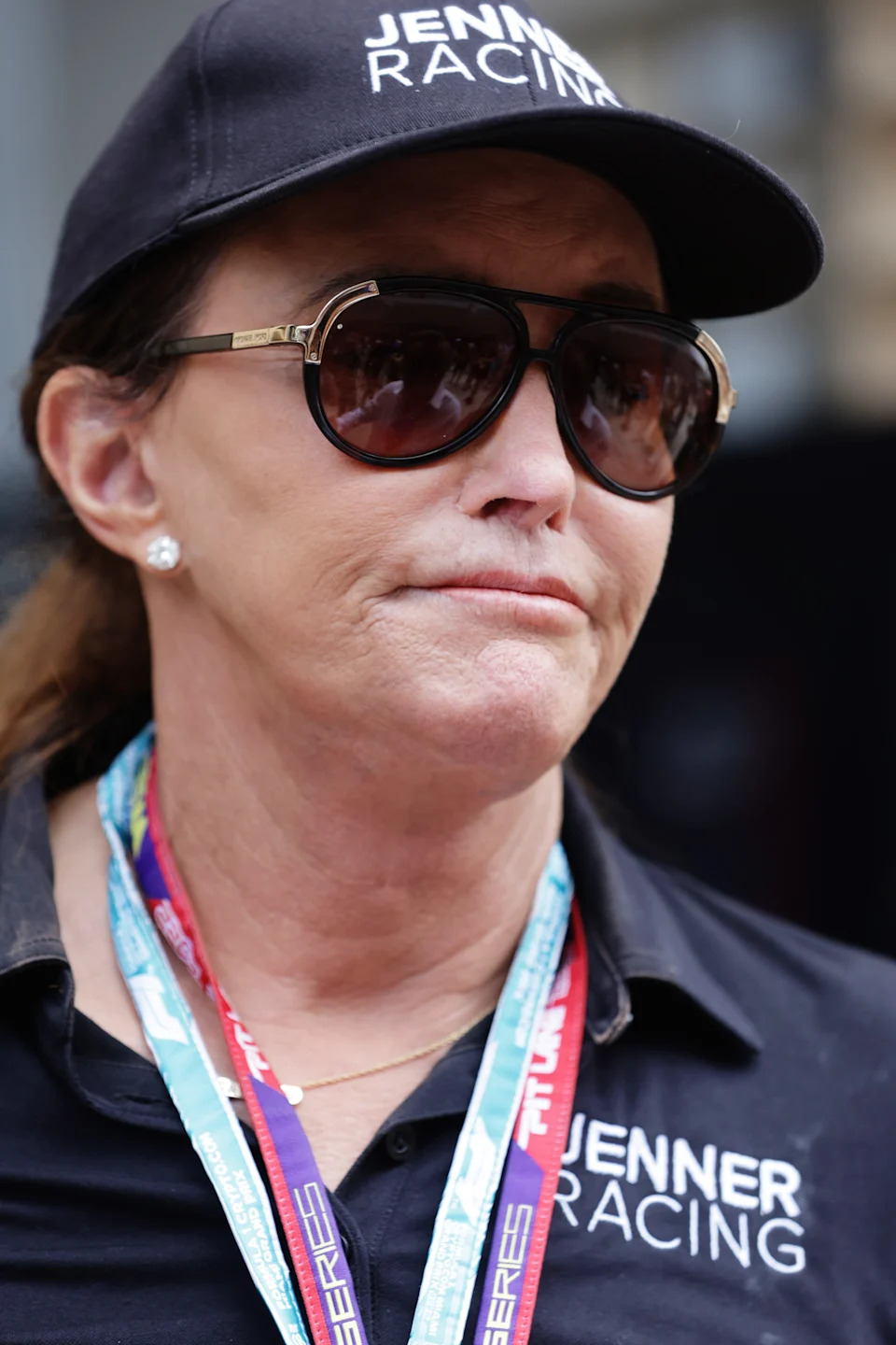 Caitlyn Jenner wearing a cap and sunglasses with branded text, attending a racing event, wearing a shirt with "Jenner Racing" text