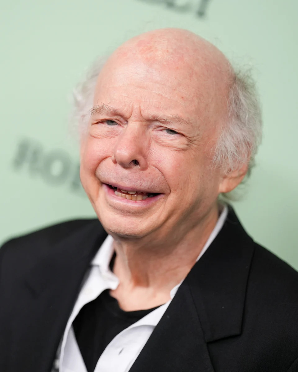 Elderly man in a suit, smiling slightly. Background blurred, suggesting an event setting
