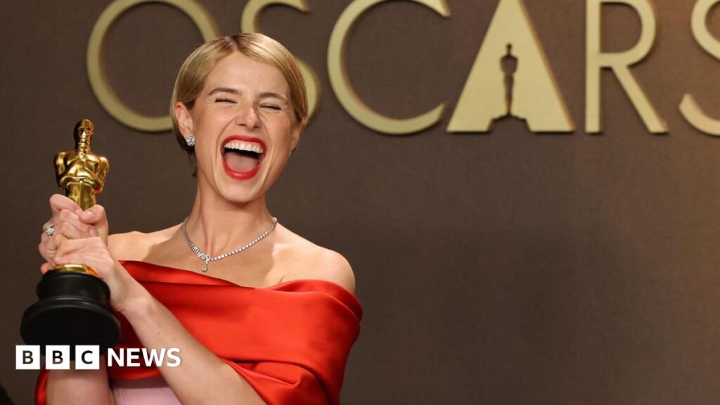 Jessie Buckley with her eyes closed smiling widely at the camera. She is holding an Oscar award and is wearing a red and pink satin dress.