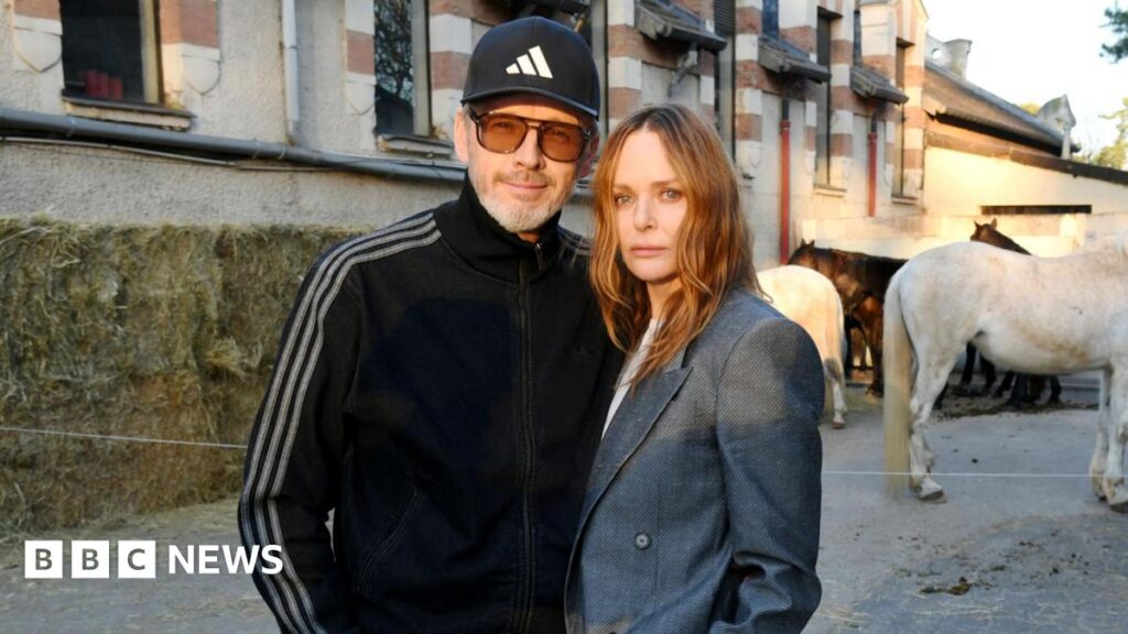 Alasdhair Willis is wearing a dark blue cap and tracksuit top and a pair of sunglasses with brown lenses. Stella McCartney has shoulder length brown hair and is wearing a grey suit jacket. The couple are standing outside a large building and there are four horses in the background.