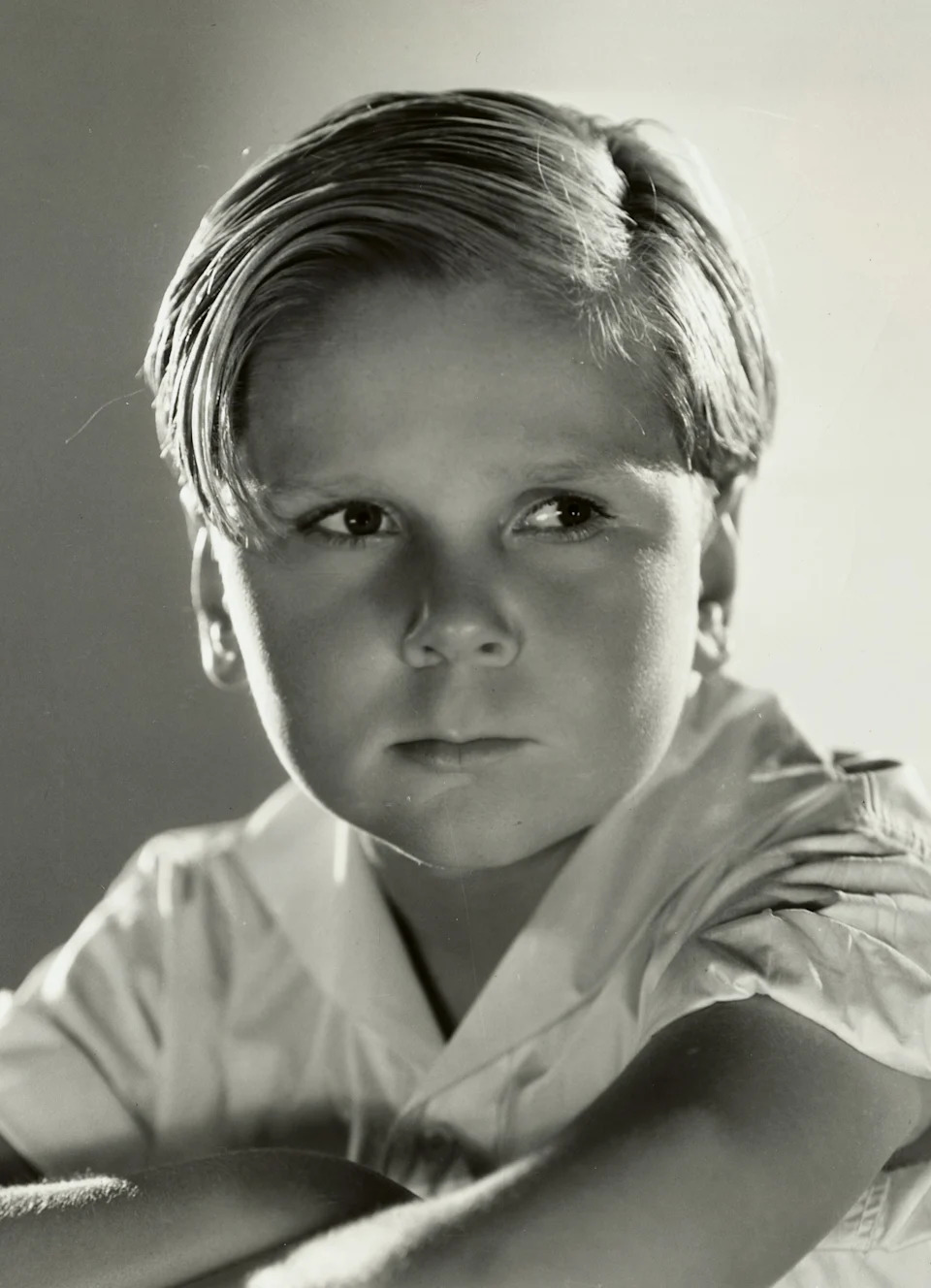 Jackie Cooper in a vintage photo poses with folded arms on a chair, looking off to the side with a pensive expression