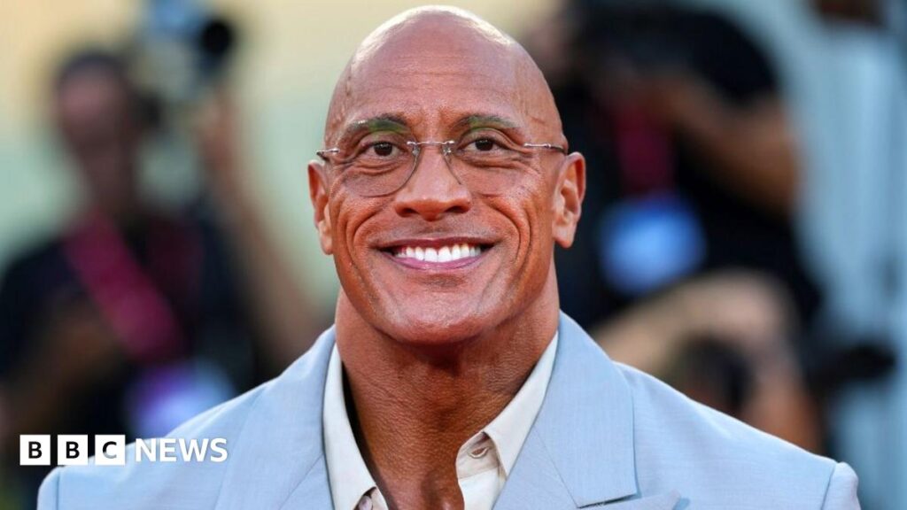 Dwayne Johnson smiles at the camera at The Smashing Machine red carpet event during last year's Venice International Film Festival. He is wearing an open-necked white shirt and a powder blue suit jacket.