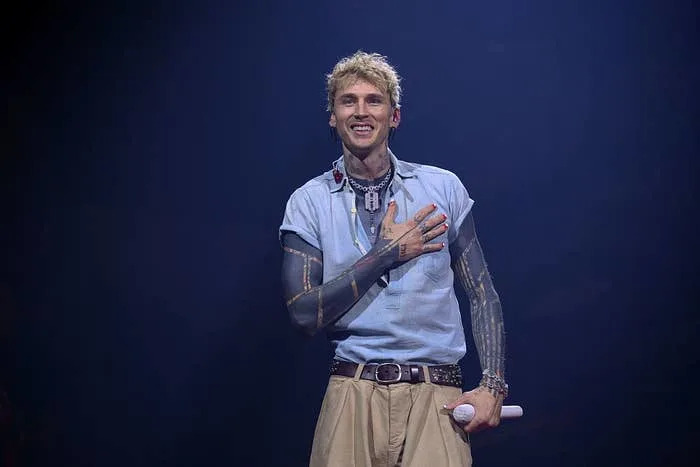 Celebrity wearing a sleeveless blue shirt and beige pants, smiling on stage while holding a microphone