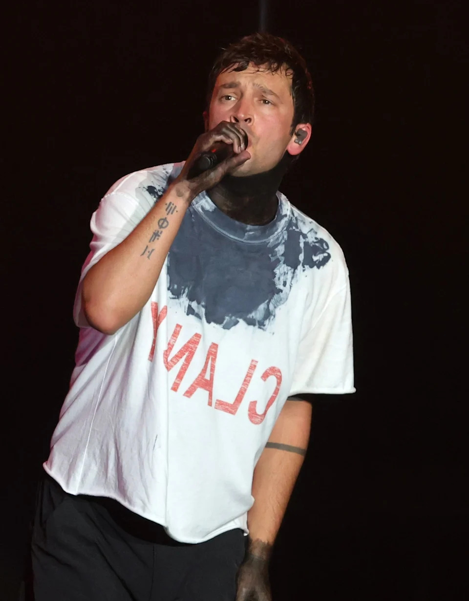 Singer performs on stage, wearing a distinctive t-shirt with a bold print, holding a microphone, engaging the audience