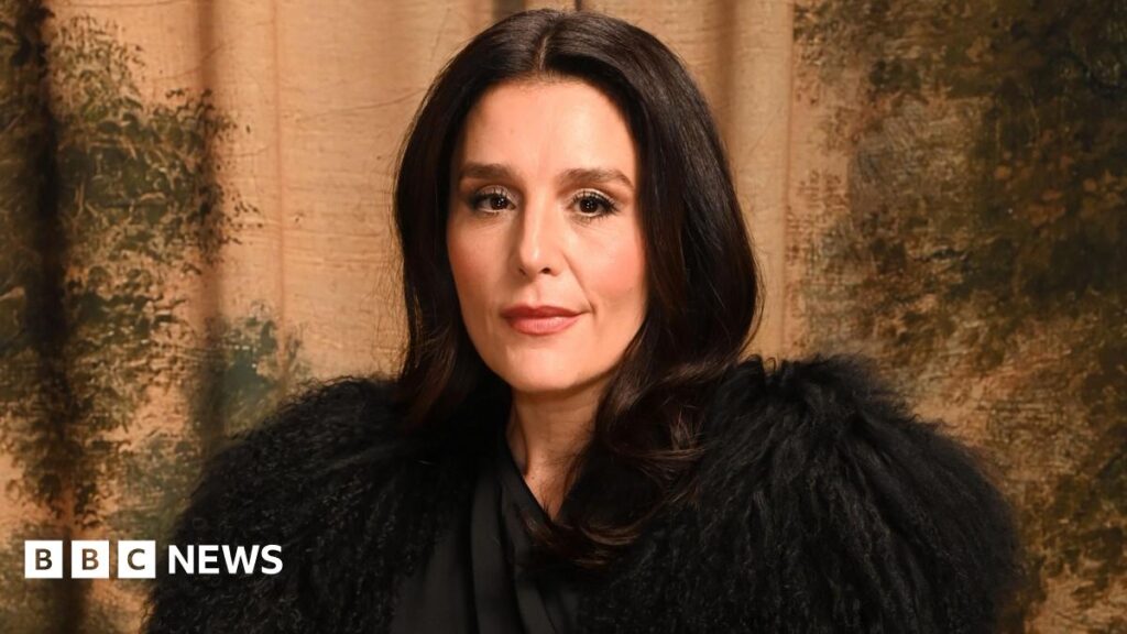 Jessie Ware poses for a photographer. She has a slight smile on her face. She is wearing a dramatic black outfit, covering her shoulders with feathery fabric. She has long, dark hair and is in front of a patterned backdrop of earthy colours.