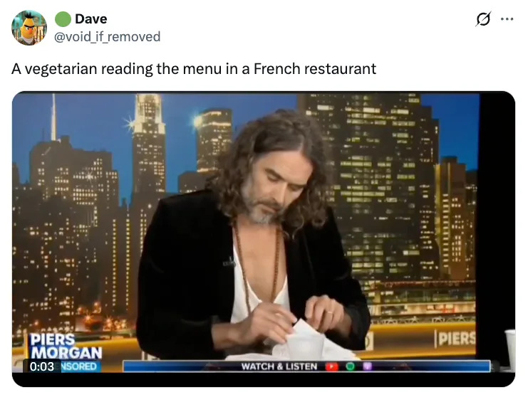 Man with long hair, wearing an open-collar shirt and velvet blazer, appears focused on a paper in a newsroom setting
