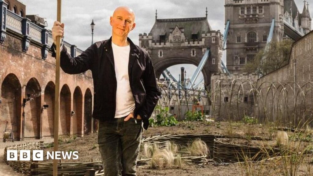 Landscape gardener and horticulturalist Nigel Dunnett wearing black jacket and trousers and white t-shirt, holding a stick with the Tower of London behind. Dunnett designed the wildflower project for Superbloom in the Tower of London moat, (date).
