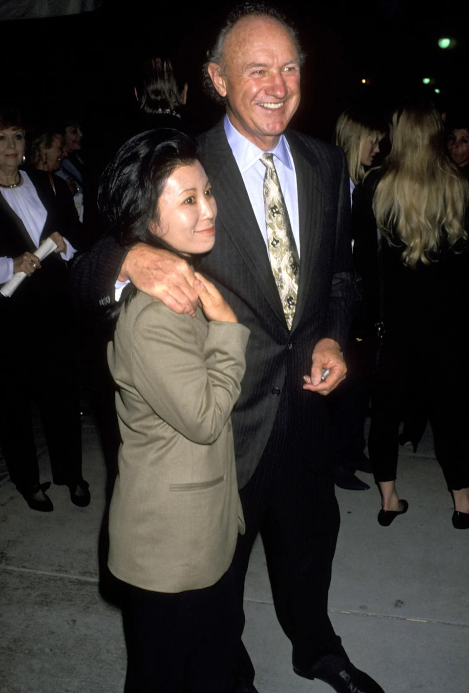 Gene Hackman in a suit with patterned tie and Betsy Arakawa in a blazer smiling at an event, surrounded by people