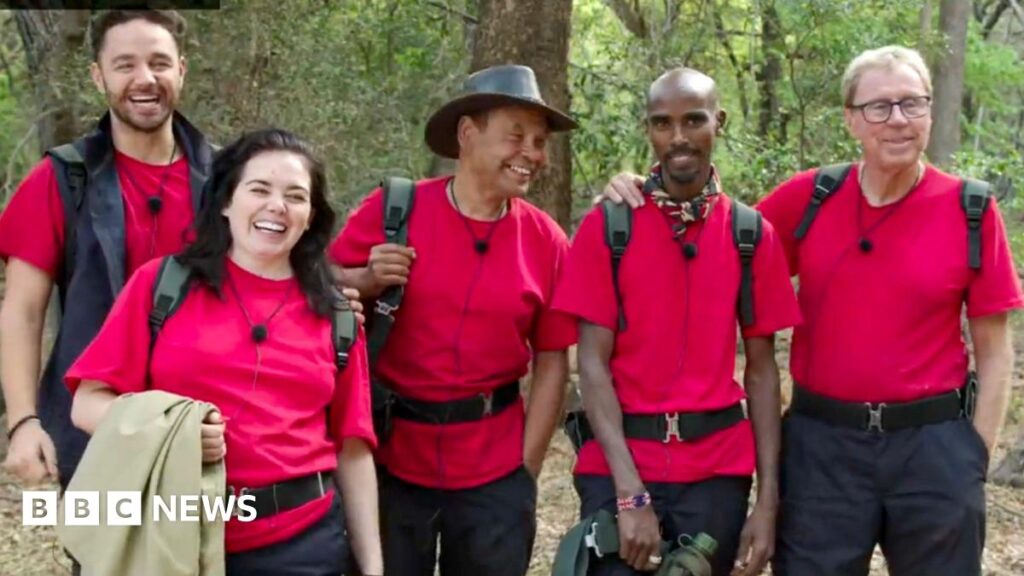 Adam Thomas, Scarlett Moffatt, Craig Charles, Mo Farah and Harry Redknapp standing together, smiling and all wearing red I'm A Celebrity T-shirts