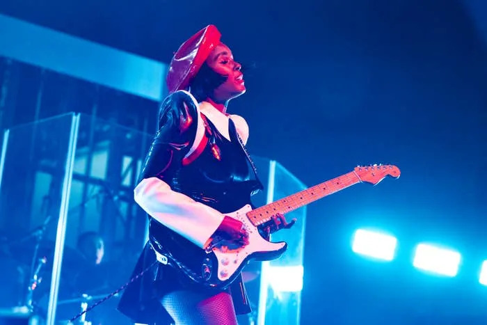 Janelle Monáe performs on stage in an edgy outfit with a coat and beret, playing an electric guitar under bright stage lights