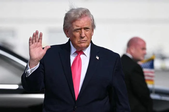 Donald Trump in a suit with a tie waves at an event, with security personnel in the background