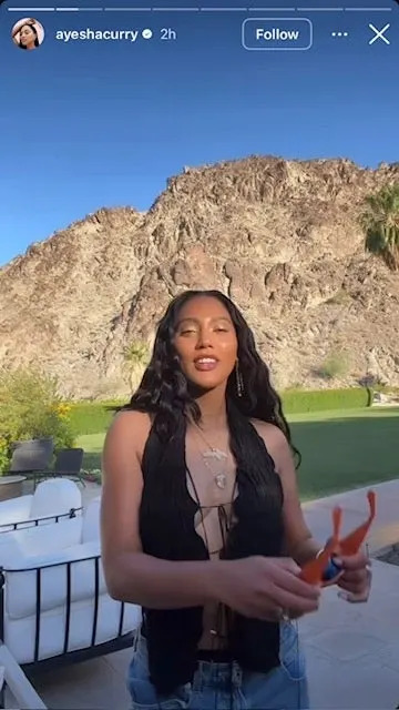 Ayesha Curry outdoors with a mountain background holds sunglasses, wearing a sleeveless top with a layered necklace and long, wavy hair