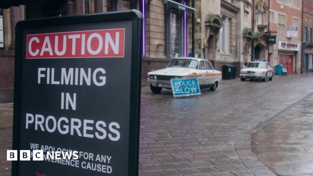 Two vintage Rover police cars, white with a red stripe across the side and blue light on top, parked up outside a row of grand-looking early 20th Century buildings, including the former Bob Carver's chip shop. A sign reads "police slow". In the foreground, another sign reads "caution, filming in progress, apologies for any inconvenience caused".