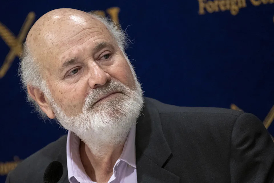 Rob Reiner with a beard turns his head at a press event, wearing a suit jacket and shirt