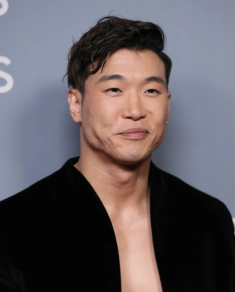 Person in a black velvet suit on a gray backdrop, smiling slightly