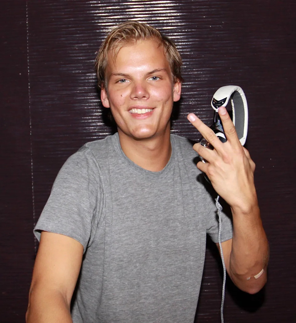 Person smiling, wearing a grey t-shirt, holds headphones in one hand with fingers forming a peace sign