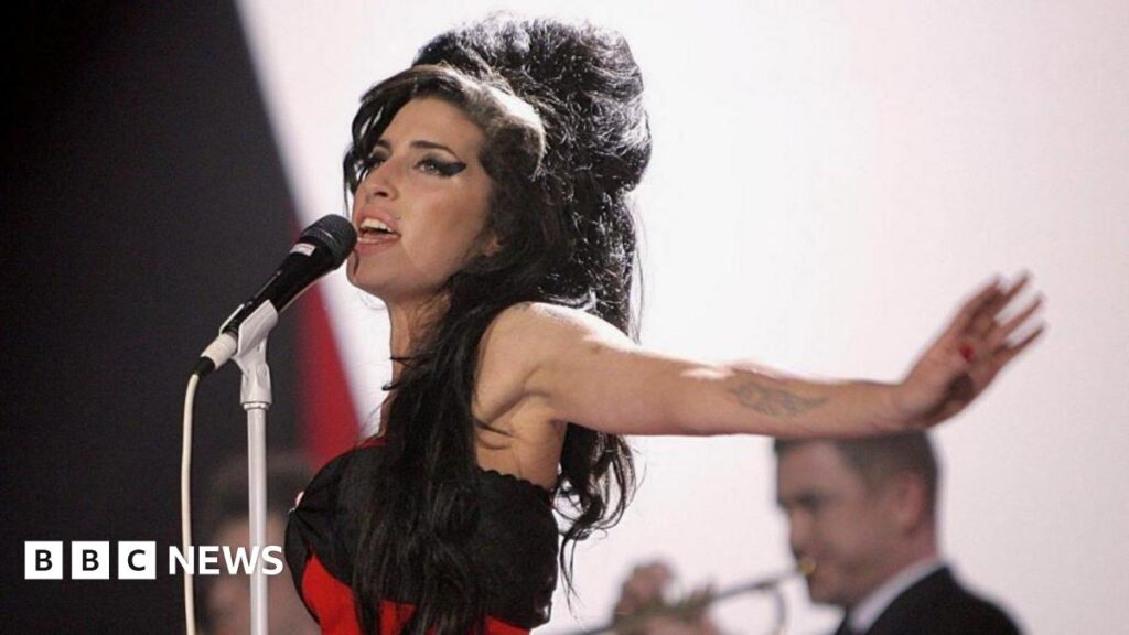 The singer with long dark hair in a messy beehive and black winged eyeliner sings into a microphone with her arms outstretched.