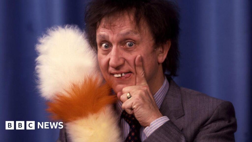 Ken stands with his trademark feather duster in hand in front of a draped curtain. His hand rests on his face. He's wearing a suit.