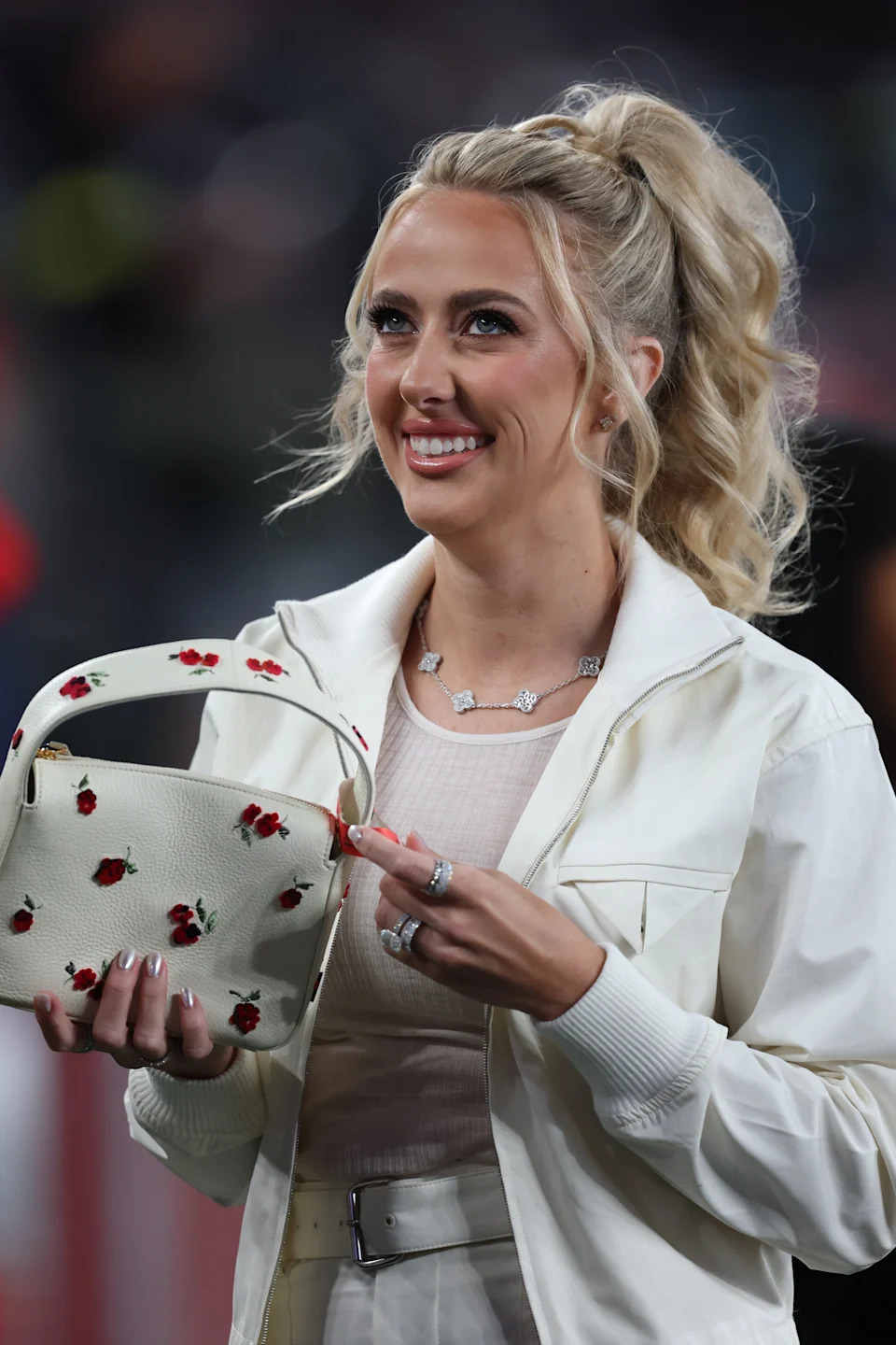Brittany Mahomes smiling while holding a floral handbag, wearing a light-colored jacket with a textured top underneath