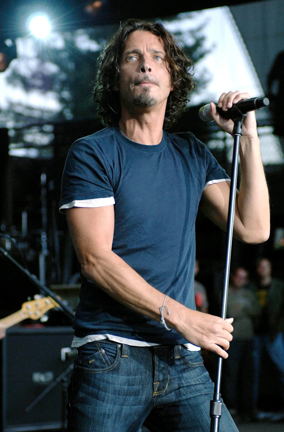 Man with curly shoulder-length hair sings into a microphone on stage, wearing a fitted t-shirt and jeans, with a focused expression